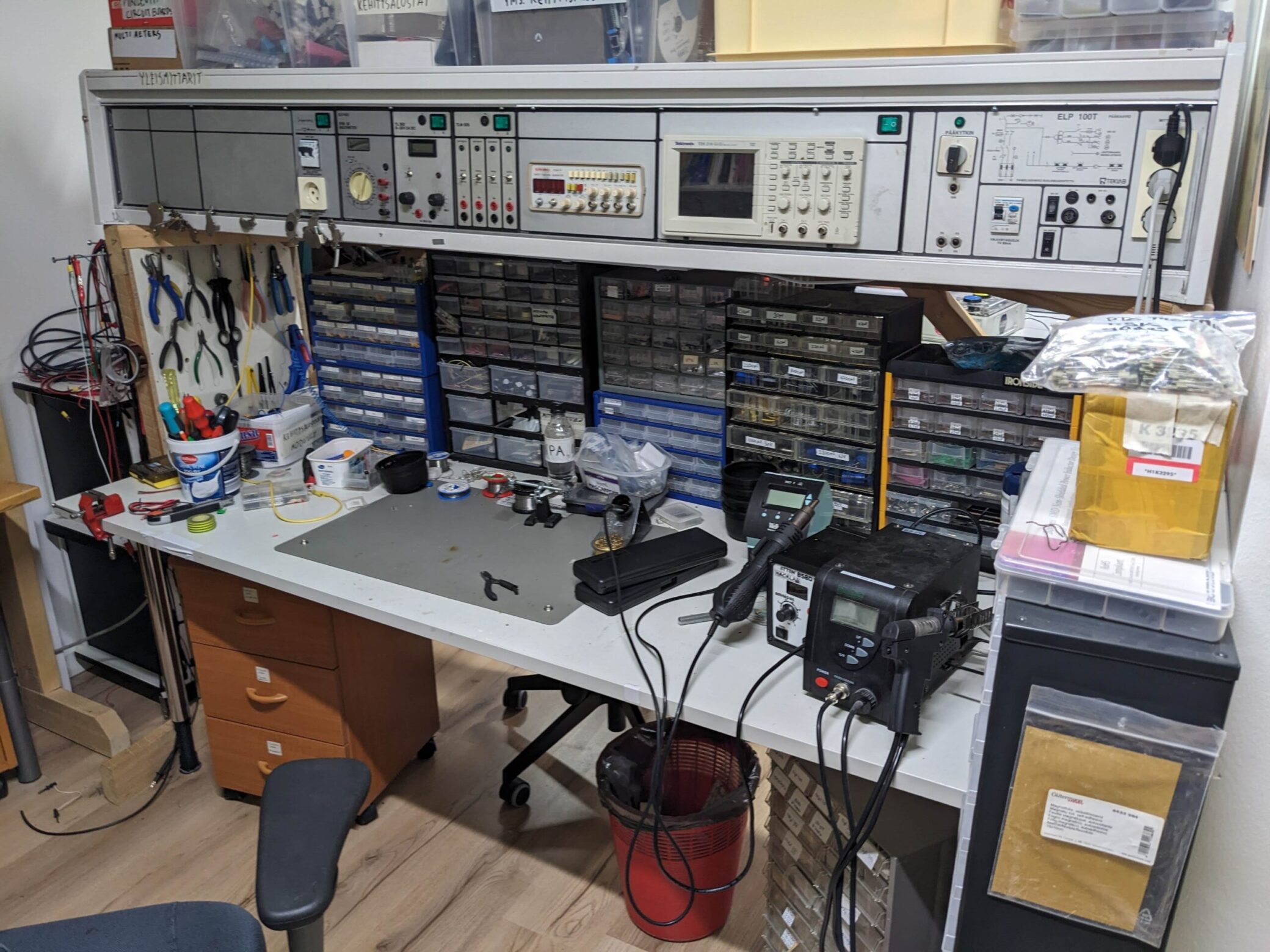 A desk featuring hand tools, soldering equipment and component drawers with electrical components, overhung by a hardware rack with buttons, displays and connection points.